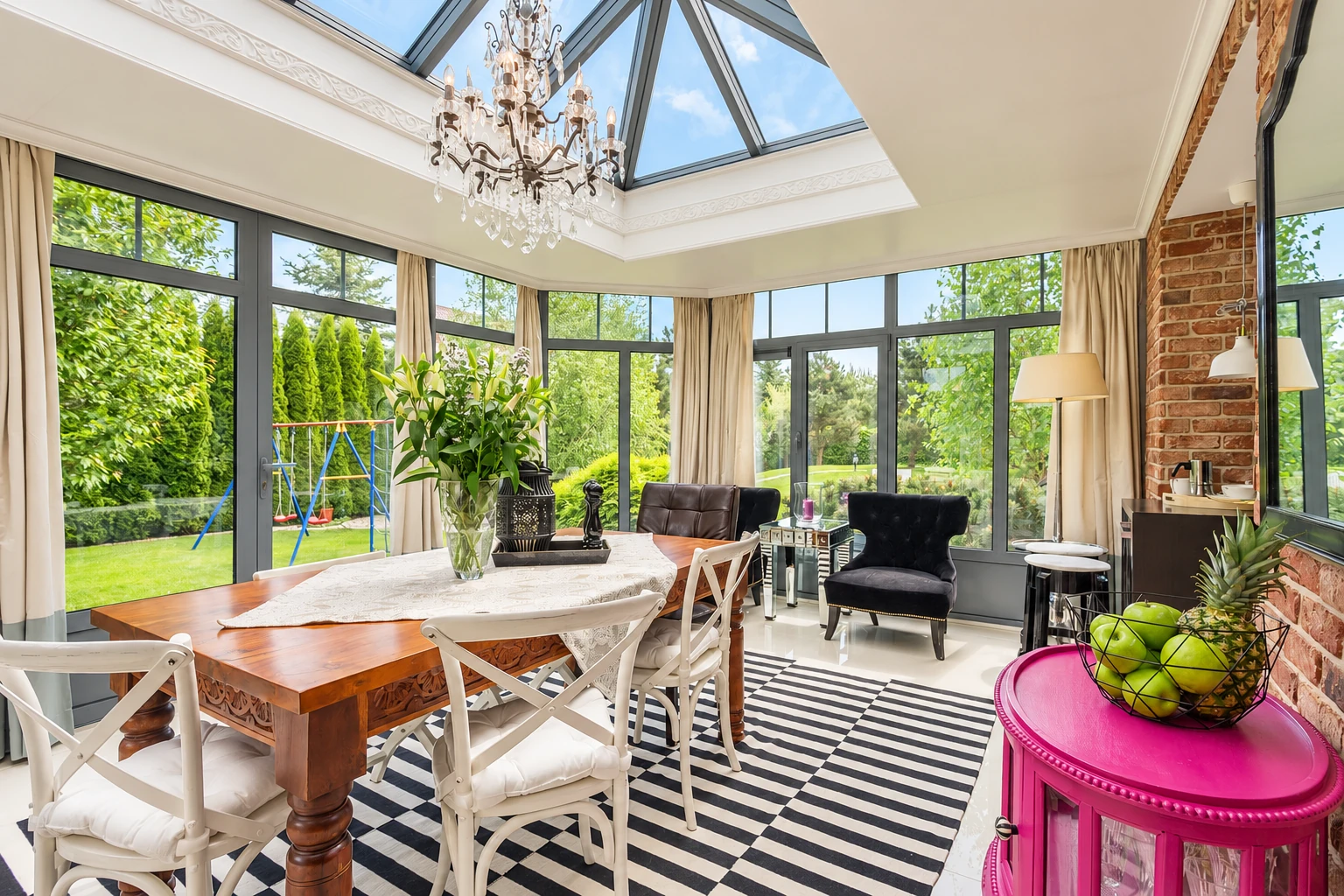 Bright four-season sunroom with a glass-paneled vaulted ceiling, chandelier, dining table, and large windows overlooking a garden.