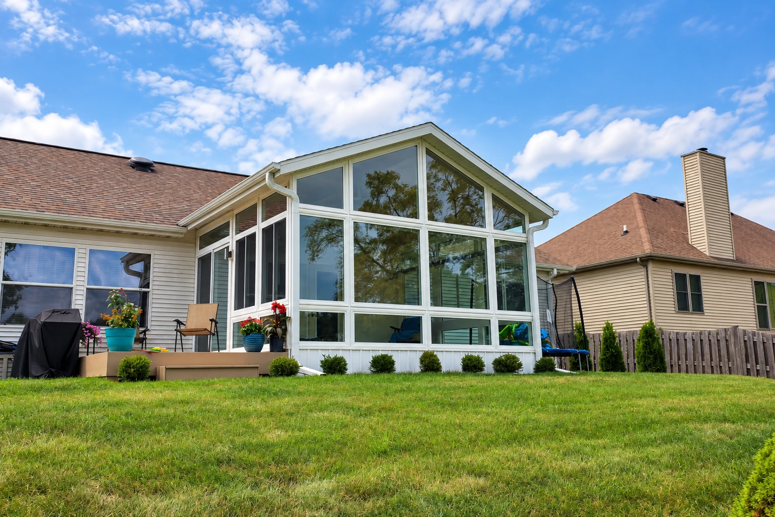 White-framed sunroom with a peaked glass roof projects from a house, overlooking a lawn and neighboring homes.