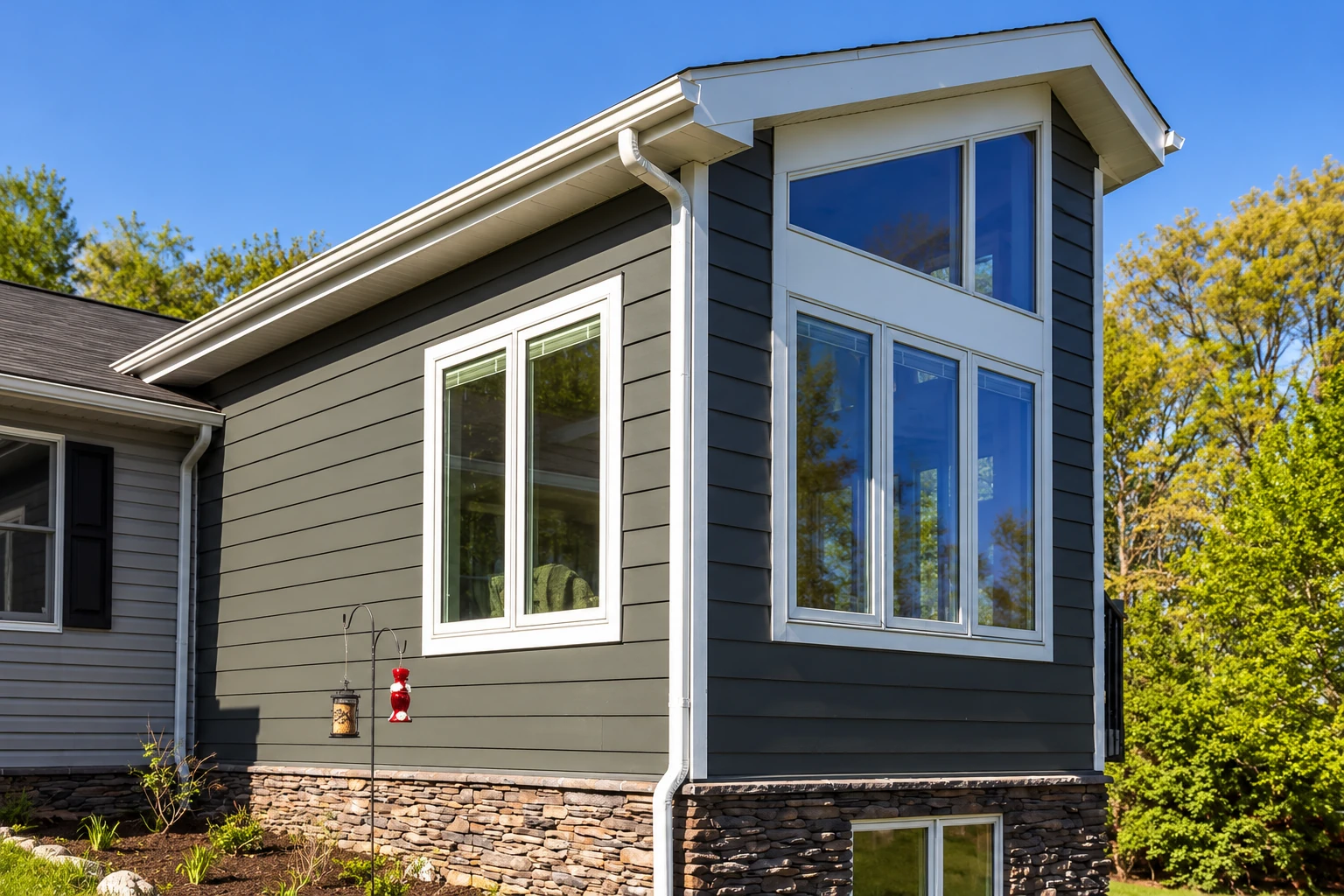 Two-story sunroom addition with dark siding, tall white-trimmed windows, and a stone foundation.