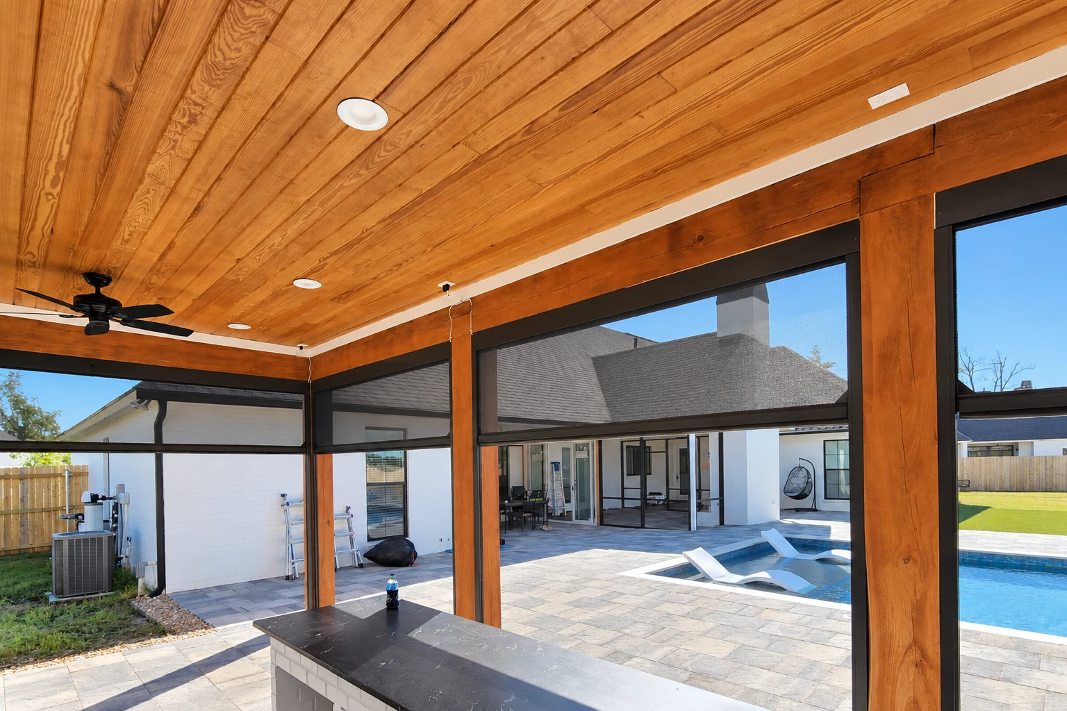 Covered patio with a warm wood-plank ceiling, ceiling fan, and dark-framed screens overlooking a pool.