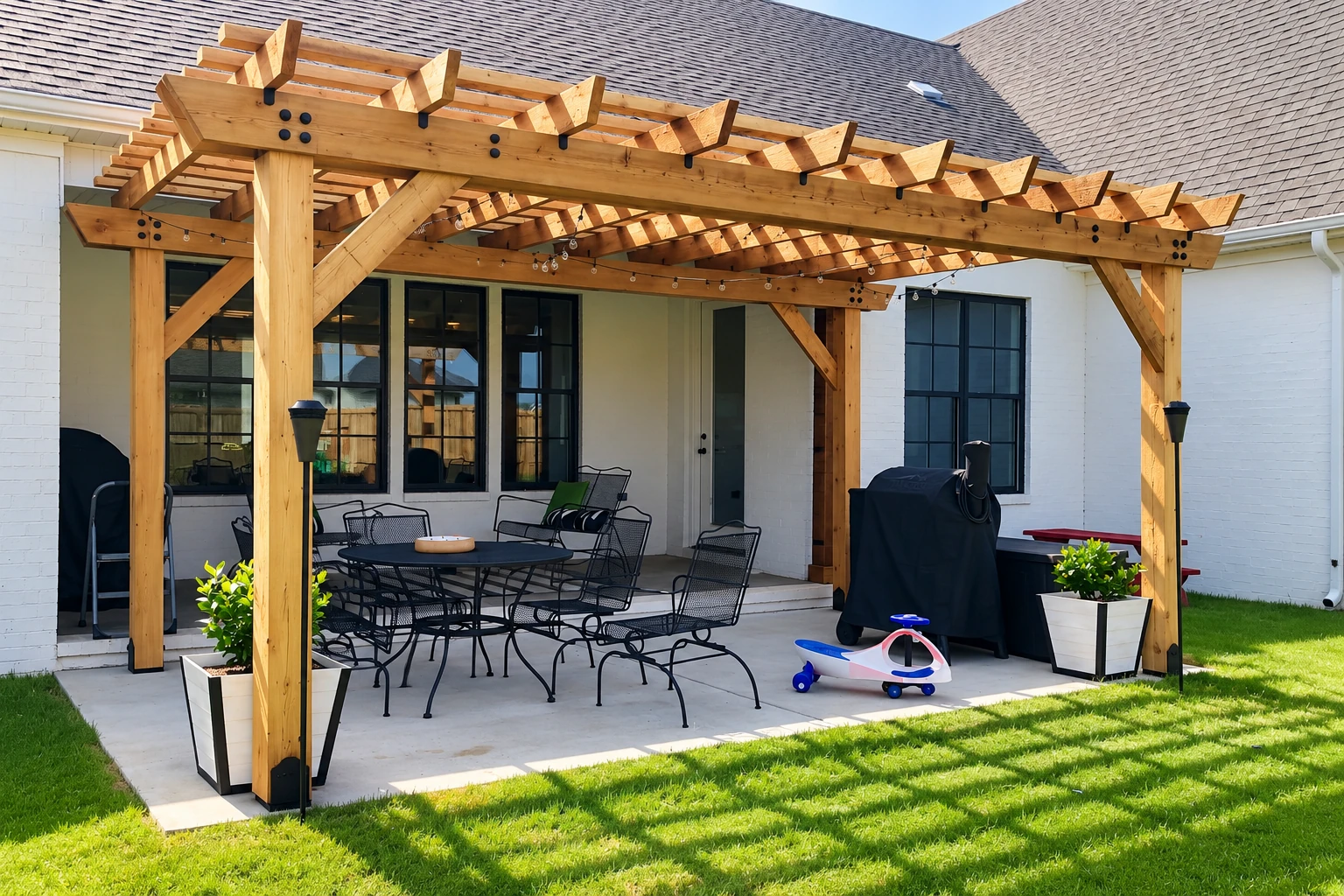Wooden patio cover with exposed beams shelters a dining set and grill beside a white house.