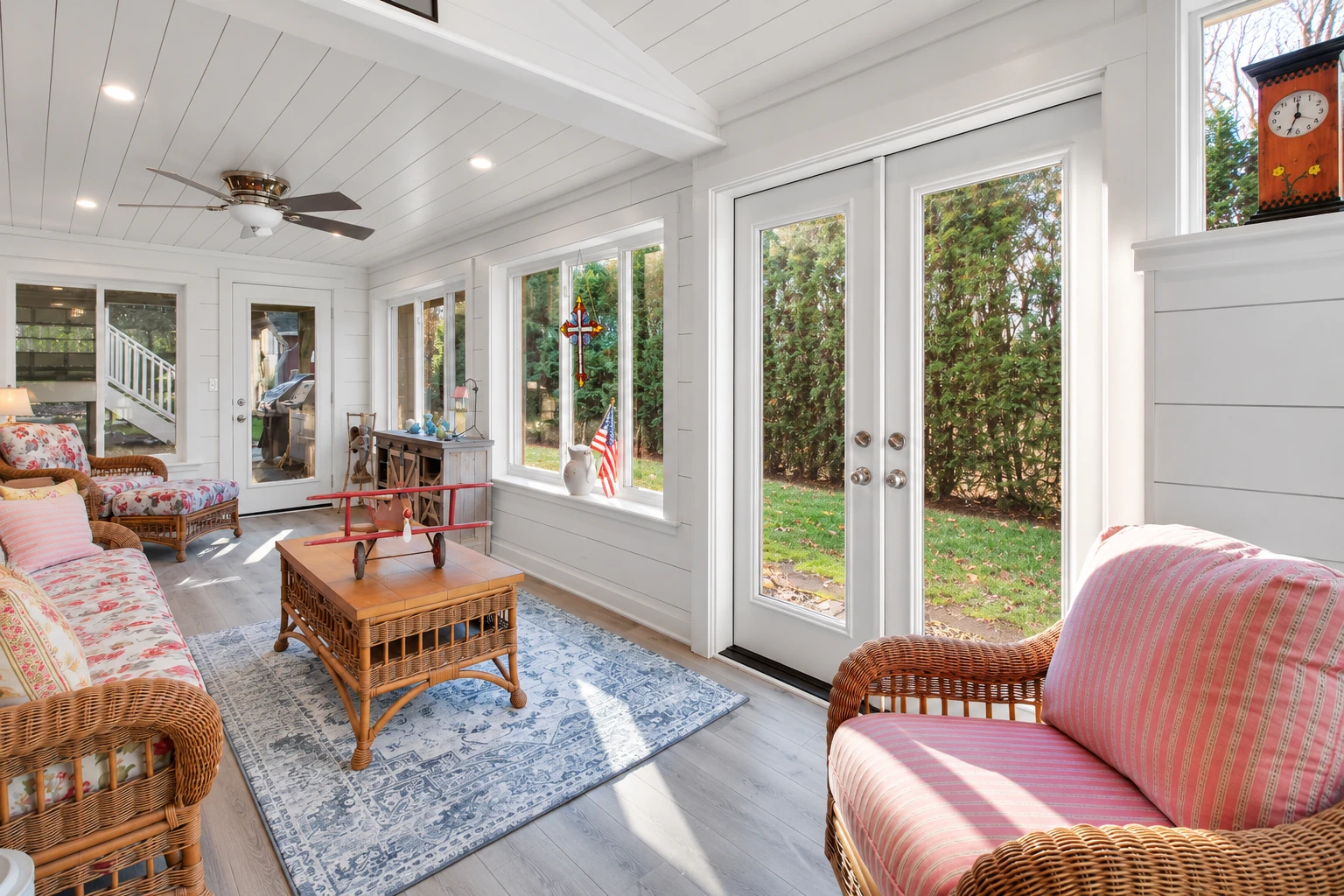 Sunroom addition with white paneled walls, wicker seating, and large glass doors and windows overlooking a garden.