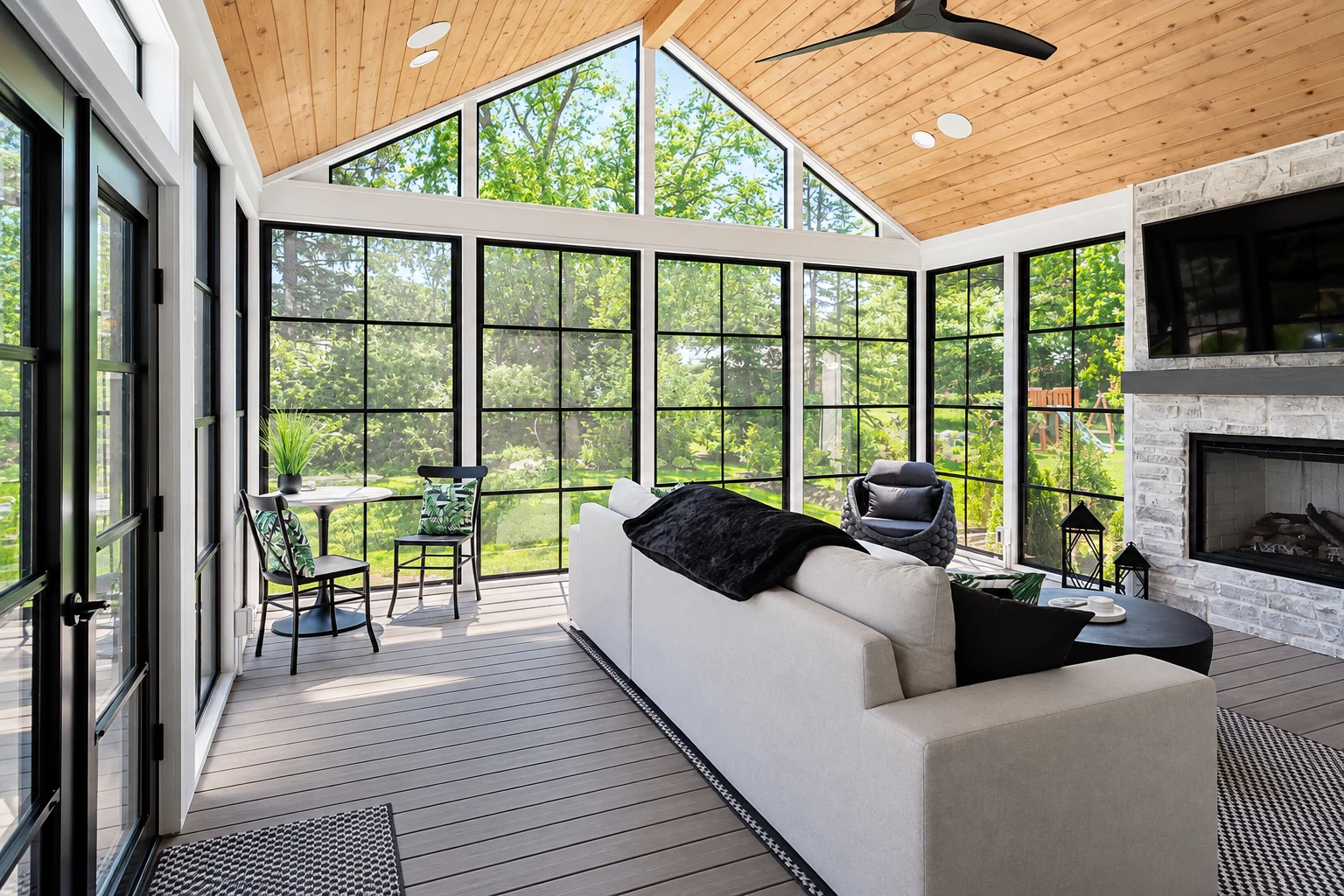 Bright sunroom with tall black-framed windows, a wood-plank vaulted ceiling, sofa, dining table and stone fireplace.