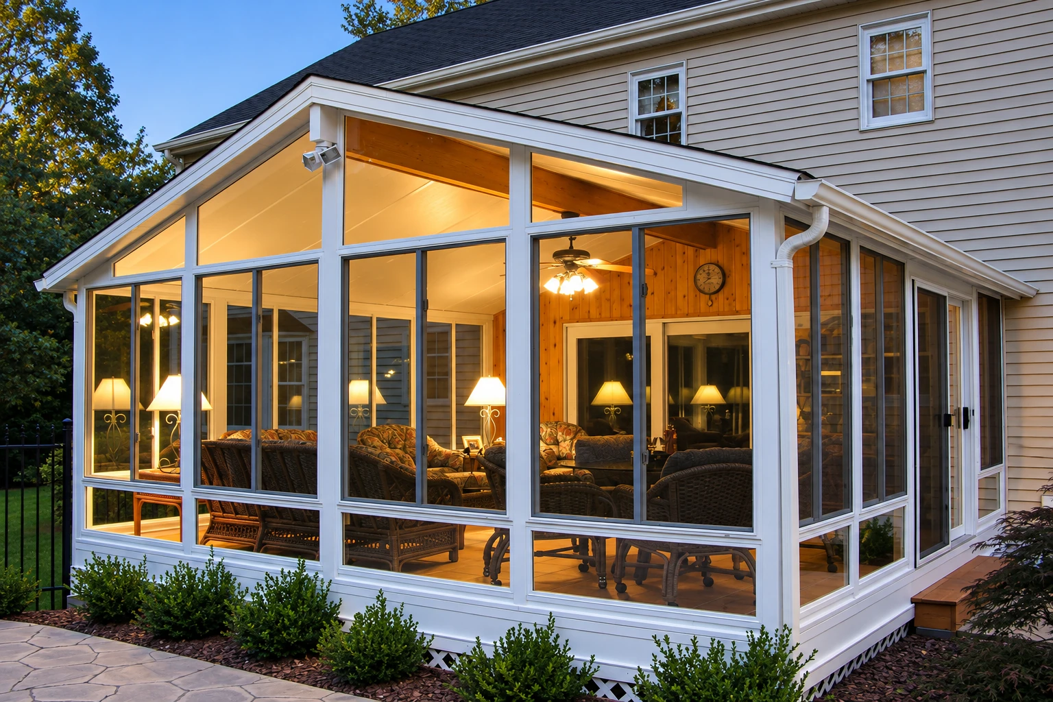 Three-season sunroom with a sloped glass-paneled roof, white framing, and warmly lit wicker seating.