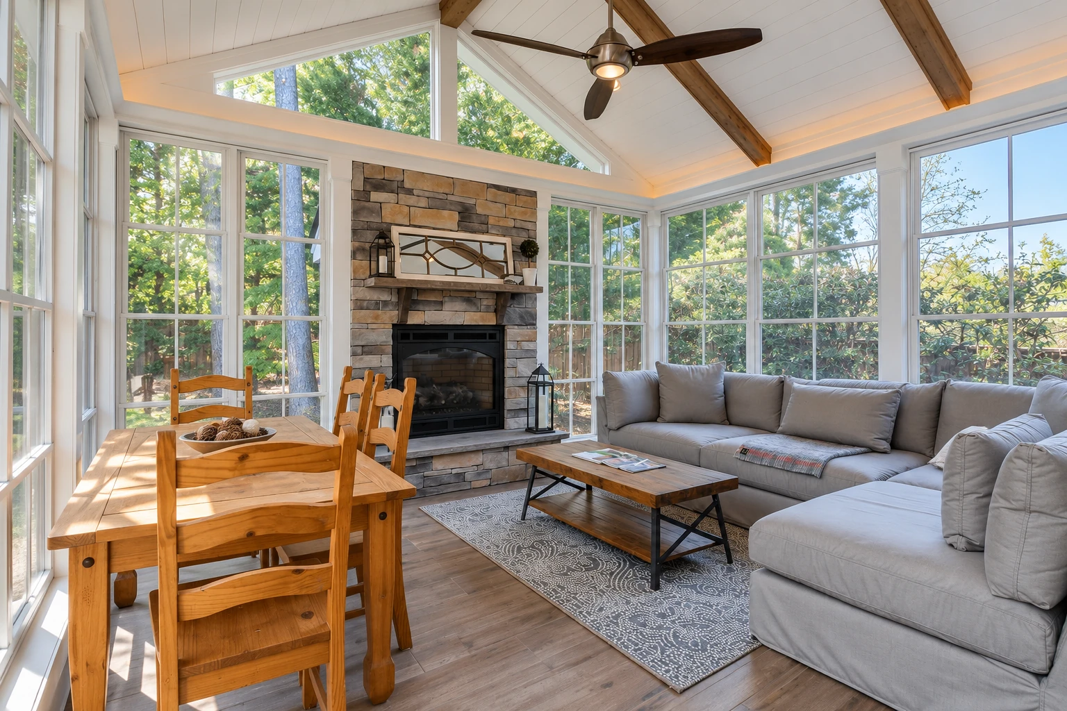 A bright three-season sunroom with large windows, a stone fireplace, dining table, ceiling fan, and gray sectional sofa.